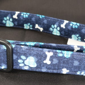 May include: A blue dog collar with a white paw print and bone pattern. The collar has a black plastic buckle.