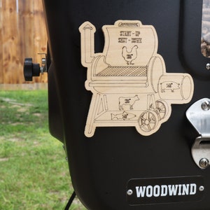 May include: A black smoker grill with a wooden sticker showing the different sizes of meat that can be cooked on the grill. The sticker says "Start-Up Meat-Week" and shows a chicken, cow, pig, and fish. The grill also has a metal bottle opener and a clear window showing wood pellets.
