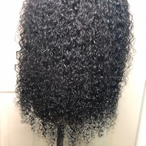 May include: A close-up of a black, curly wig. The hair is densely packed with tight, springy curls, creating a voluminous appearance. The wig is styled on a mannequin head, showcasing the texture and shape of the hair.