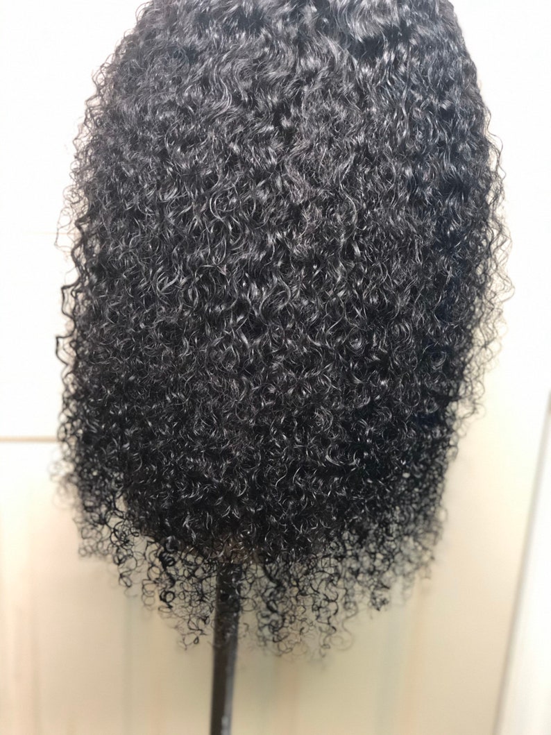 May include: A close-up of a black, curly wig. The hair is densely packed with tight, springy curls, creating a voluminous appearance. The wig is styled on a mannequin head, showcasing the texture and shape of the hair.