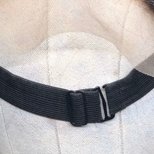 May include: Close-up of a black elastic wig band with adjustable metal clasps. The band is attached to a sheer lace front, designed to secure a wig. The background is a light-coloured fabric.