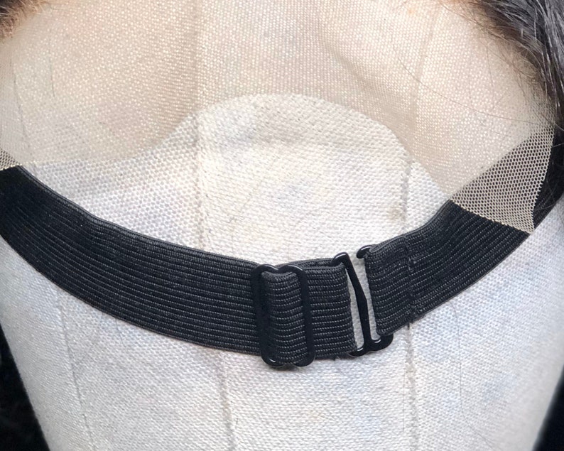 May include: Close-up of a black elastic wig band with adjustable metal clasps. The band is attached to a sheer lace front, designed to secure a wig. The background is a light-coloured fabric.