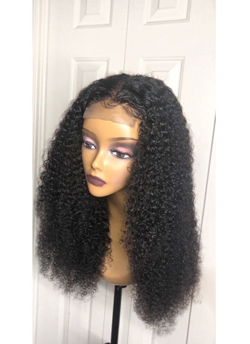 May include: A black curly wig displayed on a mannequin head. The wig has a lace front and a realistic hairline. The hair is styled in a voluminous, bouncy, curly pattern. The mannequin has a neutral skin tone and is mounted on a black stand.