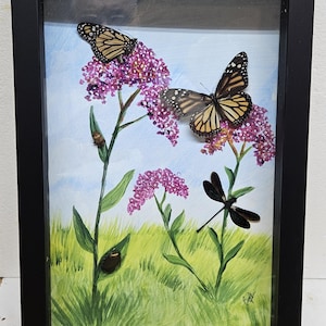 May include: A framed shadow box display featuring a painted landscape of green grass and a light blue sky. Two orange and black monarch butterflies and a black dragonfly are mounted on the surface, positioned near painted purple flower clusters.