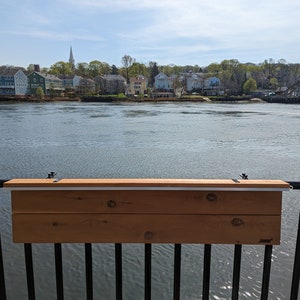 Op de afbeelding: Een houten tafel gemonteerd aan een zwart metalen hekwerk met uitzicht op een waterpartij. De tafel is gemaakt van twee houten planken en heeft een lichtbruine kleur. De tafel is met twee metalen beugels aan het hekwerk bevestigd.