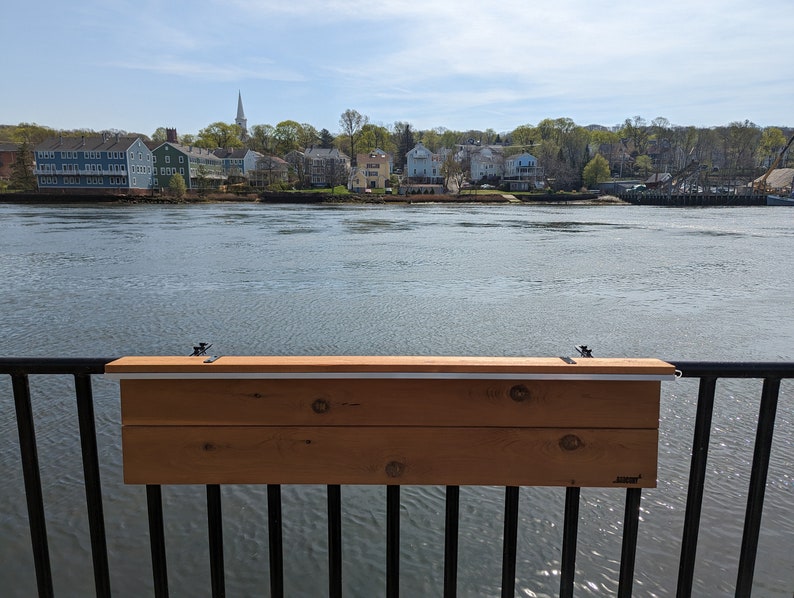 Op de afbeelding: Een houten tafel gemonteerd aan een zwart metalen hekwerk met uitzicht op een waterpartij. De tafel is gemaakt van twee houten planken en heeft een lichtbruine kleur. De tafel is met twee metalen beugels aan het hekwerk bevestigd.