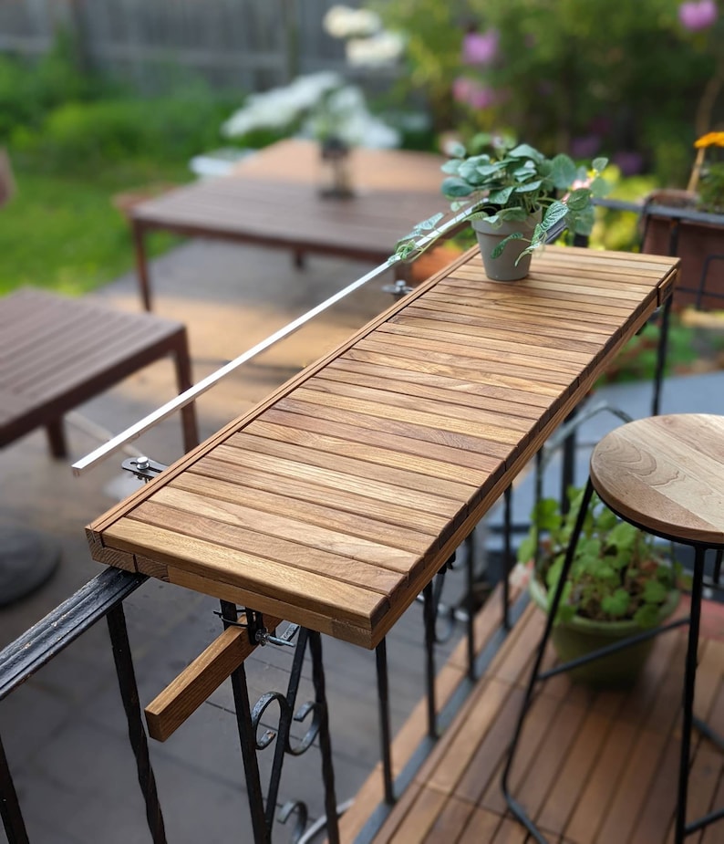 Pode incluir: Uma mesa de varanda em madeira com um tampo retangular feito de ripas de madeira estreitas e paralelas. A mesa &eacute; fixada a um corrim&atilde;o de metal preto com suportes ajust&aacute;veis. Uma pequena planta em vaso est&aacute; sobre a mesa. A mesa tem cerca de 76 cm de comprimento.