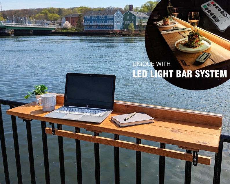 Op de afbeelding: Een houten balkontafel met een laptop, een kopje en een notitieboek erop. De tafel is bevestigd aan een zwart hekwerk en steekt uit over een waterpartij. De tafel heeft een ingebouwde LED-lichtbalksysteem met een afstandsbediening. "UNIQUE WITH LED LIGHT BAR SYSTEM" staat op de afbeelding.