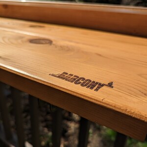Op de afbeelding: Een houten balkontafel met het woord "BALCONY" gegraveerd op het oppervlak.