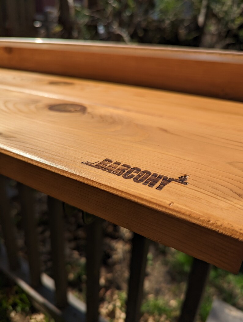 Op de afbeelding: Een houten balkontafel met het woord "BALCONY" gegraveerd op het oppervlak.