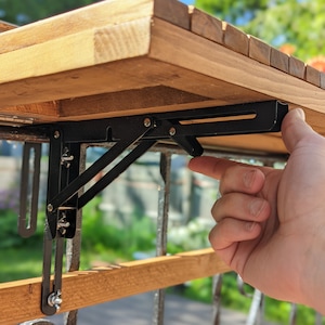 Pode incluir: Uma mesa de madeira com um suporte dobr&aacute;vel de metal preto. A mesa tem um acabamento em madeira natural e &eacute; suportada por um suporte de metal preto que se dobra para armazenamento. O suporte &eacute; fixado a um corrim&atilde;o de madeira.