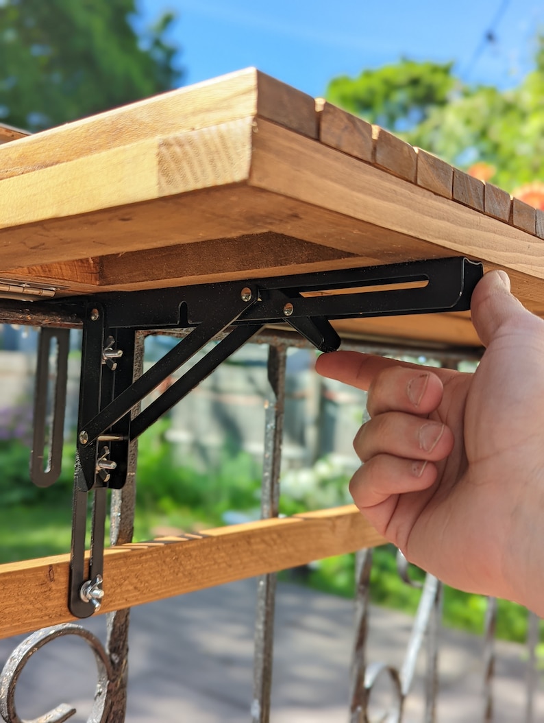 Pode incluir: Uma mesa de madeira com um suporte dobr&aacute;vel de metal preto. A mesa tem um acabamento em madeira natural e &eacute; suportada por um suporte de metal preto que se dobra para armazenamento. O suporte &eacute; fixado a um corrim&atilde;o de madeira.