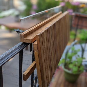 Pode incluir: Uma mesa de varanda em madeira com acabamento natural, fixada a uma grade de metal preta. A mesa &eacute; feita de ripas de madeira verticais e dobra-se para armazenamento. Projetada para uso externo.