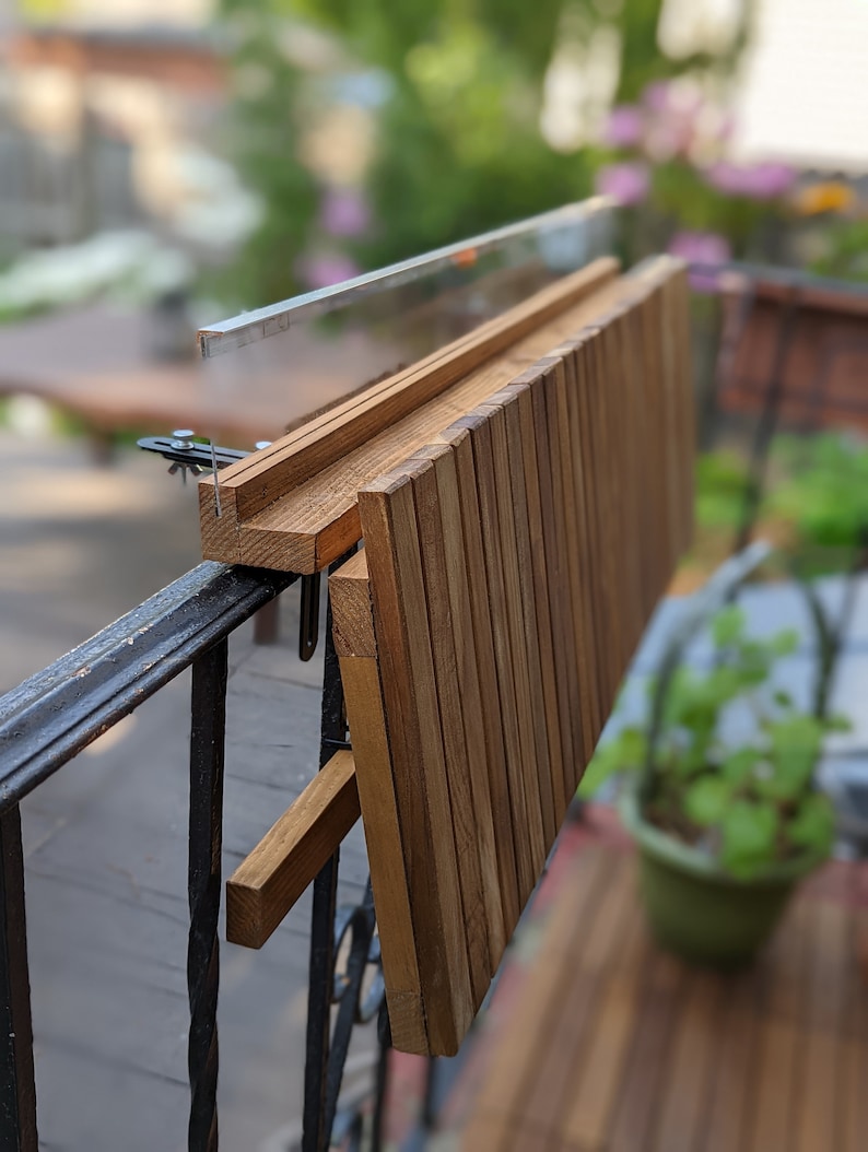 Pode incluir: Uma mesa de varanda em madeira com acabamento natural, fixada a uma grade de metal preta. A mesa &eacute; feita de ripas de madeira verticais e dobra-se para armazenamento. Projetada para uso externo.