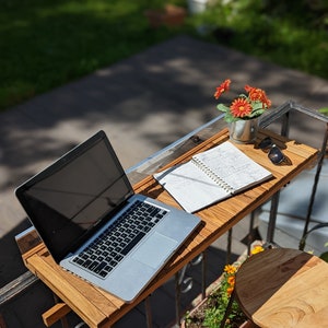 Pode incluir: Uma mesa de varanda de madeira com um laptop, um caderno, &oacute;culos de sol e um pequeno vaso com flores laranja. A mesa est&aacute; presa a uma grade de metal. Uma pequena banqueta de madeira e um vaso de flores tamb&eacute;m s&atilde;o vis&iacute;veis.
