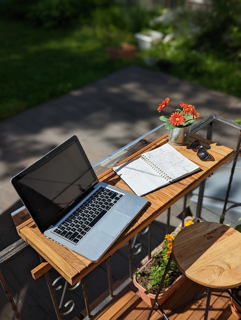 Pode incluir: Uma mesa de varanda de madeira com um laptop, um caderno, &oacute;culos de sol e um pequeno vaso com flores laranja. A mesa est&aacute; presa a uma grade de metal. Uma pequena banqueta de madeira e um vaso de flores tamb&eacute;m s&atilde;o vis&iacute;veis.