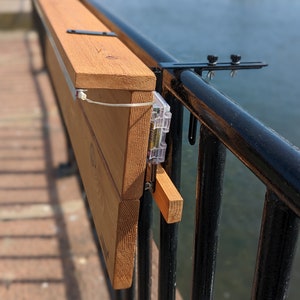 Op de afbeelding: Een houten tafel bevestigd aan een zwart metalen hek met een klem. De tafel is gemaakt van lichtbruin hout en heeft een lichtbruine vlek. De tafel is bevestigd aan het hek met een zwarte metalen klem.