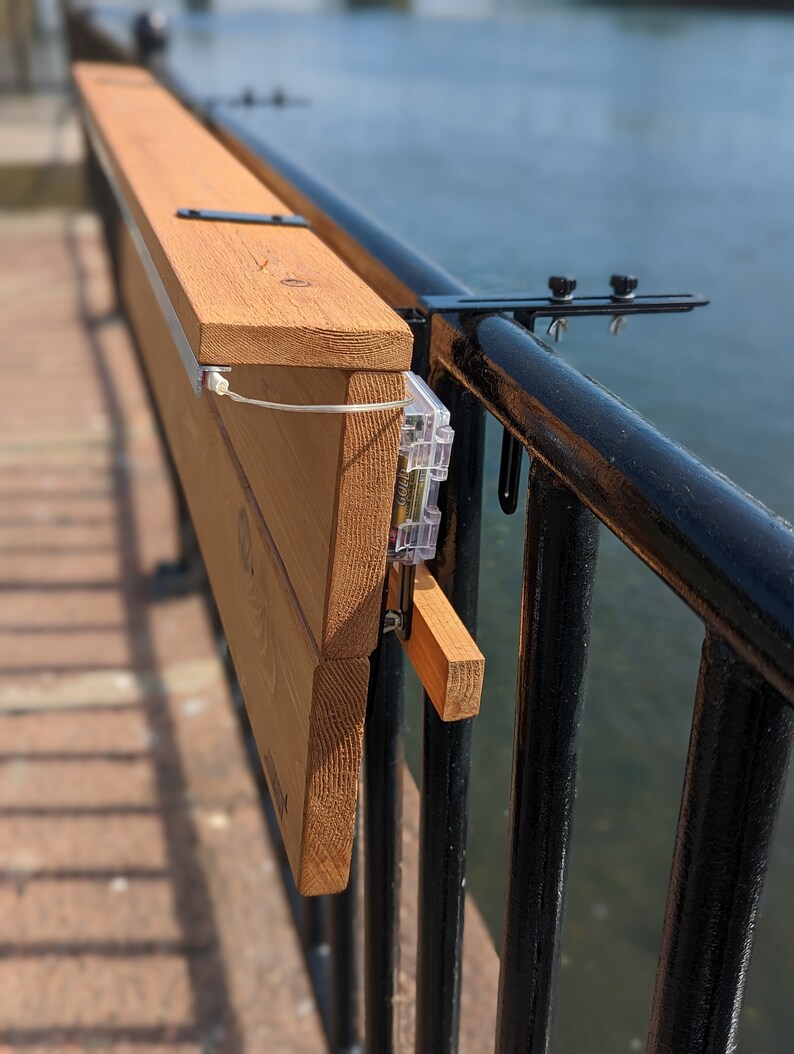 Op de afbeelding: Een houten tafel bevestigd aan een zwart metalen hek met een klem. De tafel is gemaakt van lichtbruin hout en heeft een lichtbruine vlek. De tafel is bevestigd aan het hek met een zwarte metalen klem.