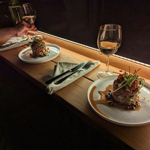 Op de afbeelding: Twee borden met eten op een houten tafel met twee glazen wijn. Het ene bord heeft een salade met een witte dressing en het andere bord heeft een steak tartare met een donkere saus.