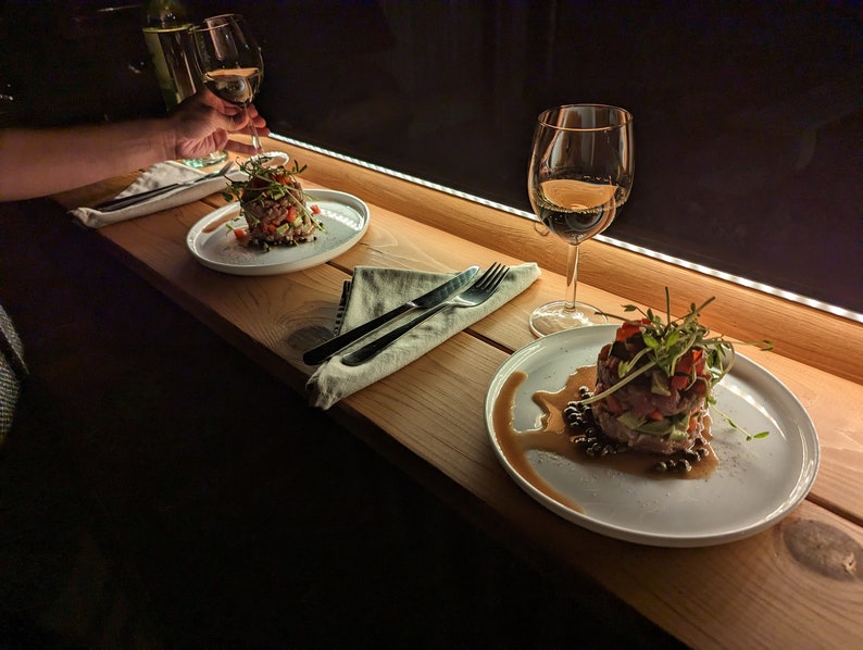 Op de afbeelding: Twee borden met eten op een houten tafel met twee glazen wijn. Het ene bord heeft een salade met een witte dressing en het andere bord heeft een steak tartare met een donkere saus.