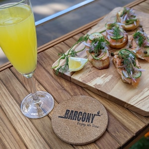 Pode incluir: Um copo de mimosa ao lado de uma t&aacute;bua de madeira com aperitivos. A t&aacute;bua cont&eacute;m p&atilde;o torrado coberto com salm&atilde;o, pepino, cebola roxa e endro. Uma base de corti&ccedil;a com o texto "BARCONY Enjoy the View" est&aacute; sobre a mesa de madeira.