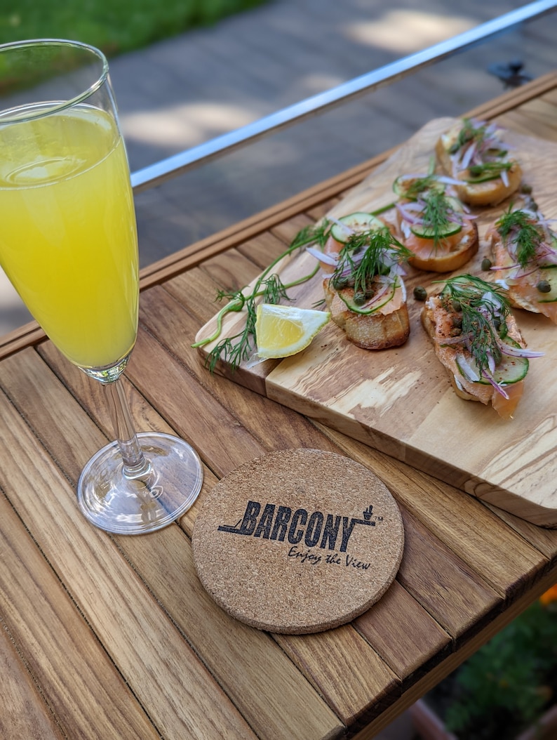 Pode incluir: Um copo de mimosa ao lado de uma t&aacute;bua de madeira com aperitivos. A t&aacute;bua cont&eacute;m p&atilde;o torrado coberto com salm&atilde;o, pepino, cebola roxa e endro. Uma base de corti&ccedil;a com o texto "BARCONY Enjoy the View" est&aacute; sobre a mesa de madeira.