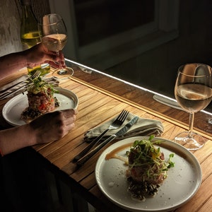 Pode incluir: Uma cena de jantar com pouca luz com dois pratos de comida, cada um guarnecido com microvegetais. Cada prato est&aacute; sobre uma mesa de madeira, acompanhado por talheres e uma toalha de linho. Ta&ccedil;as de vinho cheias de vinho branco.