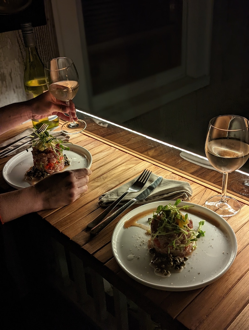 Pode incluir: Uma cena de jantar com pouca luz com dois pratos de comida, cada um guarnecido com microvegetais. Cada prato est&aacute; sobre uma mesa de madeira, acompanhado por talheres e uma toalha de linho. Ta&ccedil;as de vinho cheias de vinho branco.
