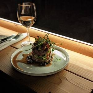 Op de afbeelding: Een wit bord met een gelaagde salade bedekt met spruiten en een donkere saus. Het bord staat op een houten tafel met een glas witte wijn op de achtergrond.