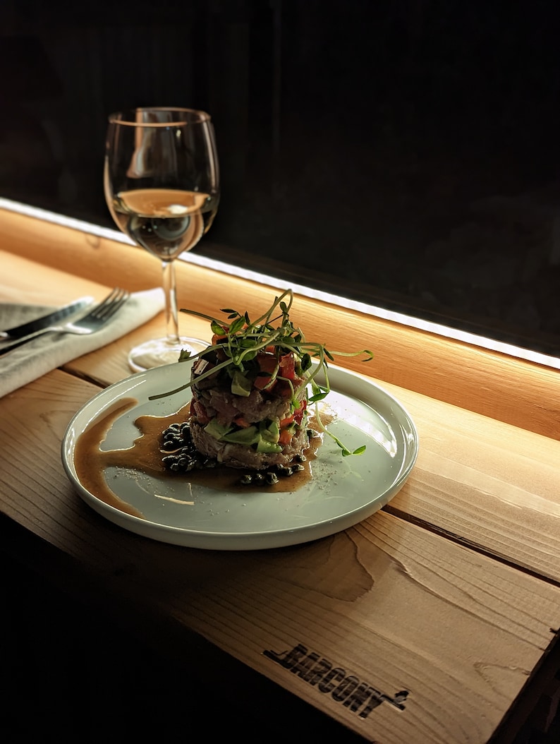 Op de afbeelding: Een wit bord met een gelaagde salade bedekt met spruiten en een donkere saus. Het bord staat op een houten tafel met een glas witte wijn op de achtergrond.