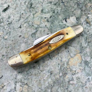 May include: A closed Case pocket knife with a brown and cream handle and silver-coloured bolsters. The knife has a single blade and the Case logo is visible. It rests on a speckled grey surface.