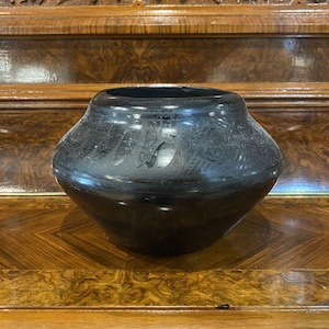 May include: A black, handcrafted pottery bowl with a wide, rounded shape. The bowl has a smooth, glossy finish and sits on a wooden surface. The dark color and simple design give it a modern, yet rustic feel. The bowl is approximately 6 inches in diameter.