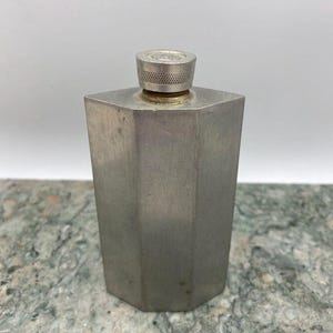 May include: A silver-colored, hexagonal flask with a screw-on cap. The flask has a brushed metal finish and a geometric design. The cap is a smaller, circular piece with a textured edge. The flask is sitting on a mottled green and gray surface.
