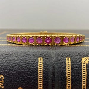 May include: A gold-colored bangle bracelet with a row of pink gemstones. The bracelet is set with multiple round, pink gemstones. The bracelet is resting on a black surface.