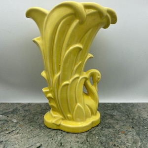 May include: A yellow ceramic vase with a swan design. The vase has a curved, organic shape with stylized leaves and a swan silhouette. The vase is approximately 8 inches tall.