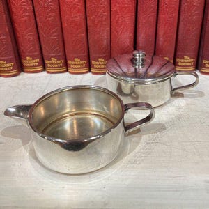 May include: Two antique silver serving pieces, a creamer with a spout and handle, and a covered sugar bowl. The items are highly reflective, with a bright, polished finish. Behind them are a row of red books with gold lettering.