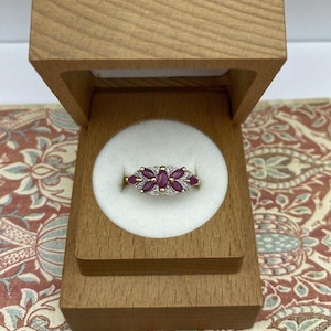 May include: A ring featuring three marquise-cut deep pink stones arranged horizontally, surrounded by small clear accent stones in a metal setting, displayed inside a light-colored wooden box with a white circular insert for the ring.
