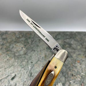 May include: A vintage pocket knife with a partially open blade. The blade is engraved with "1905 75th ANNIVERSARY 1980". The handle is a light brown colour with a cream-coloured bolster and the word "CASE" is visible on the handle.