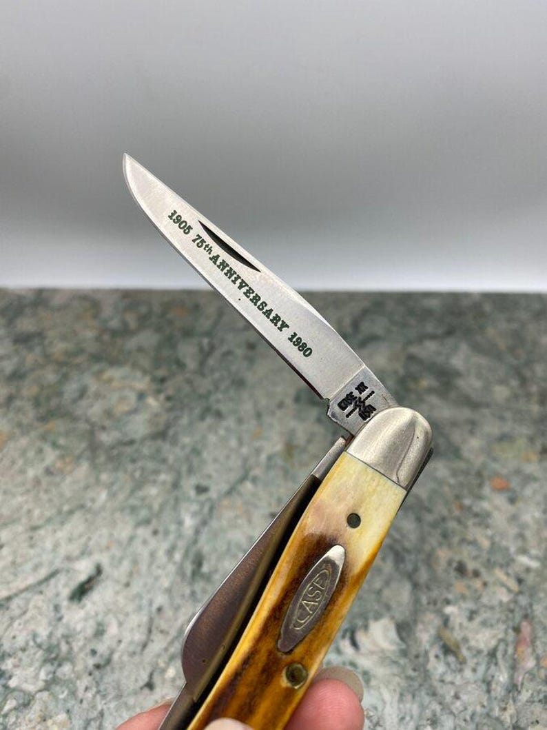 May include: A vintage pocket knife with a partially open blade. The blade is engraved with "1905 75th ANNIVERSARY 1980". The handle is a light brown colour with a cream-coloured bolster and the word "CASE" is visible on the handle.