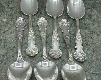 May include: Six silver spoons with ornate floral designs on the handles. The spoons have a polished, reflective surface and a classic, elegant aesthetic. The spoons are arranged on a mottled gray surface.