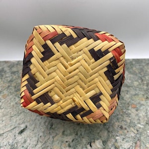 K&ouml;nnte beinhalten: Ein quadratischer Webkorb mit geometrischem Muster. Der Korb besteht aus naturfarbenen, braunen und roten Webstreifen. Der Korb steht auf einer grau-gr&uuml;nen Oberfl&auml;che.
