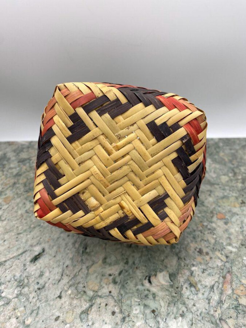 K&ouml;nnte beinhalten: Ein quadratischer Webkorb mit geometrischem Muster. Der Korb besteht aus naturfarbenen, braunen und roten Webstreifen. Der Korb steht auf einer grau-gr&uuml;nen Oberfl&auml;che.