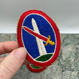 May include: A vintage military patch with a red, white, and blue design. The patch features a white obelisk with a red sword and a yellow handle. The patch is surrounded by a red border with a green base.