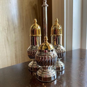 May include: A silver and gold-toned condiment set, including two shakers and a central container with a tall, decorative handle. The set features a ribbed design on the lower portion and a gold-colored, ornate top. The items are displayed on a dark wooden surface.