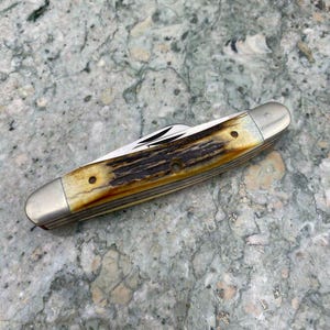 May include: A closed pocket knife with a bone handle and silver-coloured metal ends. The handle has a brown and cream colour pattern. The knife blades are visible. The knife is lying on a grey and green speckled surface.