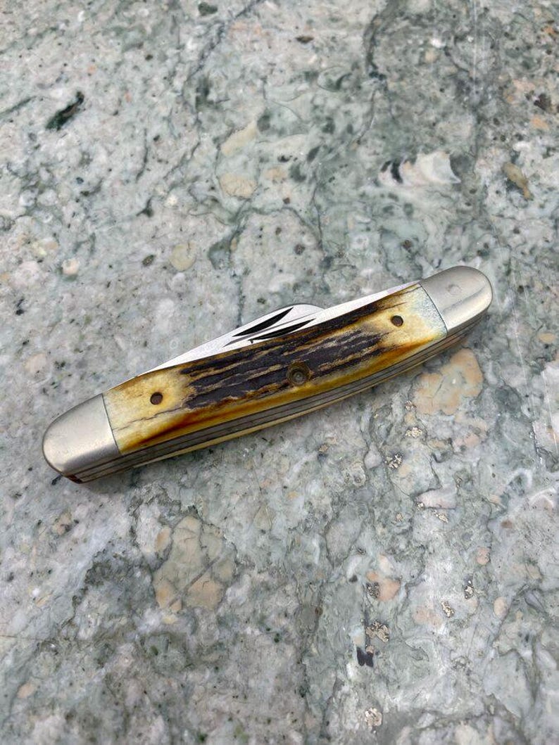 May include: A closed pocket knife with a bone handle and silver-coloured metal ends. The handle has a brown and cream colour pattern. The knife blades are visible. The knife is lying on a grey and green speckled surface.