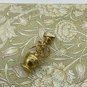 May include: A small, gold-colored hookah with intricate detailing. The miniature hookah features a bulbous base, a slender stem, and a coiled hose. The design includes etched patterns and a small mouthpiece. The hookah is set against a floral patterned background.