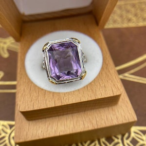 May include: A rectangular amethyst gemstone ring with gold and silver accents, set in a wooden jewelry box. The ring's purple stone is the focal point, with a detailed metalwork setting.