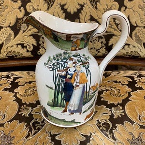 May include: White ceramic pitcher with a curved handle and spout. The pitcher features a colorful illustration of two people in period clothing, with trees and a building in the background. The rim and handle are trimmed with brown.