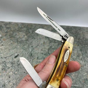 May include: A Case pocket knife with three open blades. The handle is a mix of yellow and brown, with a silver bolster and the Case logo. The blade reads "1905 75th Anniversary 1980".